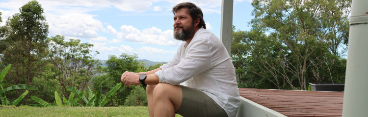 Man sitting on house steps with backyard garden and mountains in the background.